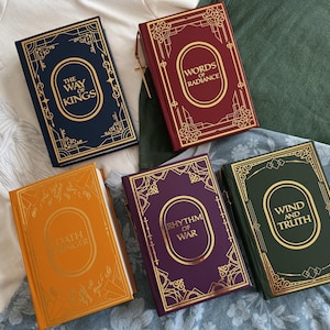 Puede incluir: Cinco libros de tapa dura con letras doradas y bordes decorativos. Los libros son de varios colores: azul marino, burdeos, naranja, morado y verde. Los títulos incluyen "The Way of Kings", "Words of Radiance", "Oathbringer", "Rhythm of War" y "Wind and Truth".