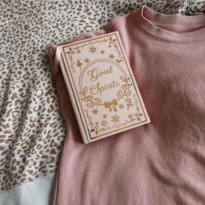 May include: A pink sweatshirt with a book that reads "Good Spirits" in gold lettering. The book has decorative gold accents. A leopard print sweatshirt is partially visible in the background.