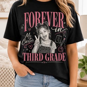 Może przedstawiać: Czarny t-shirt z krótkim rękawem z nadrukiem graficznym zawierającym tekst FOREVER in, Ms. Carter oraz THIRD GRADE. Wzór zawiera czarno-białe zdjęcie dziecka wykonującego znak pokoju, notatnik z napisem BEST DAY EVER, ołówek oraz serca.