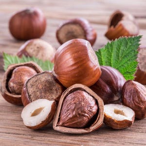 May include: A collection of whole and halved brown hazelnuts with their hard, woody shells and smooth, cream-colored kernels. Some nuts are partially cracked open, revealing the textured interior, accompanied by a bright green serrated leaf.