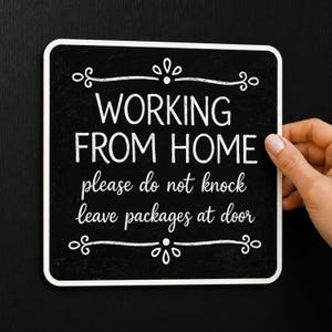 Op de afbeelding: Een vierkant zwart bord met afgeronde hoeken en witte tekst: WORKING FROM HOME, please do not knock, leave packages at door. De tekst wordt omlijst door decoratieve witte krullen aan de boven- en onderkant van het bord.