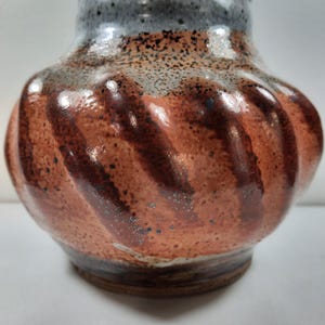 May include: A handcrafted ceramic vase with a unique, textured design. The vase features a dark brown base, transitioning to a reddish-brown body with a spiral pattern. The rim is a speckled grey, adding contrast to the earthy tones.