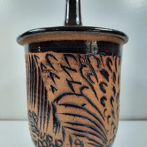 Peut inclure: Un pot en céramique fait à la main avec un couvercle. Le pot est de couleur marron chaud avec des motifs sculptés complexes et un bord et un couvercle marron foncé. Le couvercle a une petite poignée. Le pot est probablement utilisé pour le stockage.