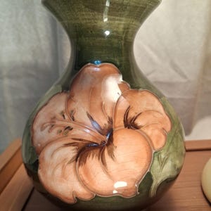 May include: A ceramic vase with a glossy dark olive green glaze and a prominent, raised relief peach-coloured hibiscus flower design on the front. The surface features a fine crackle finish throughout, with a wide neck and a rounded body.
