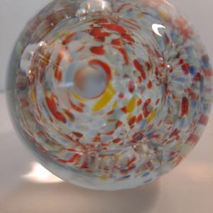 Peut inclure: Un presse-papier en verre transparent de forme sphérique. L'intérieur présente un tourbillon de taches colorées rouges, jaunes et bleues sur fond blanc. Le verre est lisse et réfléchit la lumière, créant un effet décoratif.