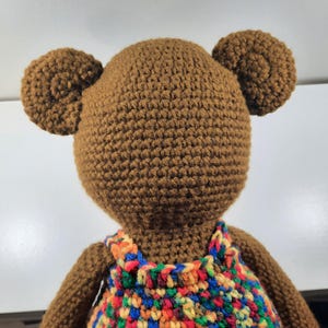 May include: A handmade brown crochet bear with two round ears. The bear is wearing a colorful crochet dress with red, blue, orange, and yellow accents. The bear is viewed from the back.