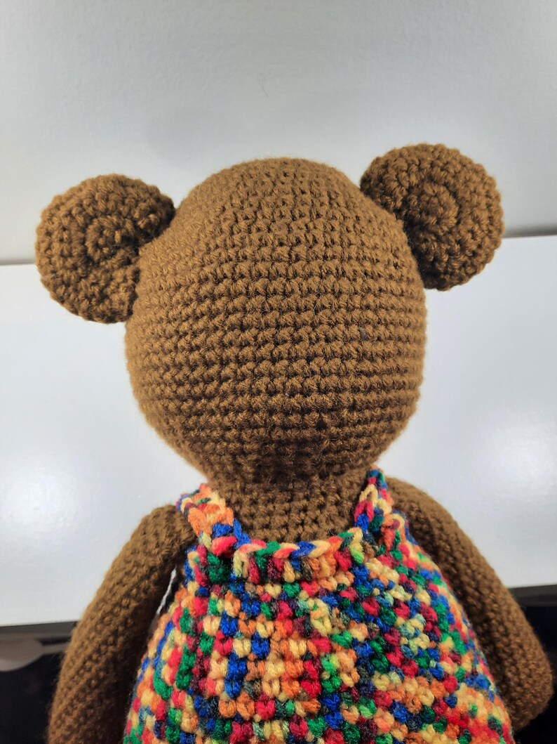May include: A handmade brown crochet bear with two round ears. The bear is wearing a colorful crochet dress with red, blue, orange, and yellow accents. The bear is viewed from the back.