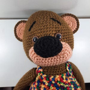 May include: Crocheted brown bear with a black nose, tan muzzle, and large black eyes. The bear wears a colorful, patterned overall. The bear's ears are tan with brown trim. The bear is a handmade item.