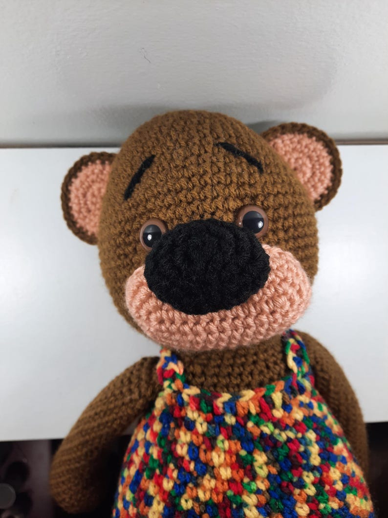 May include: Crocheted brown bear with a black nose, tan muzzle, and large black eyes. The bear wears a colorful, patterned overall. The bear's ears are tan with brown trim. The bear is a handmade item.