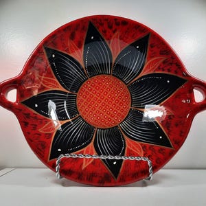 May include: A red ceramic serving dish with a black and white floral design. The dish has a sunflower pattern in the centre and handles on either side. The dish is displayed on a silver metal stand.