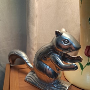 May include: A silver-colored metal nutcracker shaped like a squirrel sitting on a log. The squirrel has a textured tail, defined facial features, and a visible screw on its side. The object is constructed with a hinged mechanism for cracking nuts.