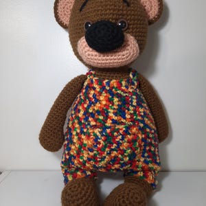 May include: A handmade brown crochet bear doll wearing colorful overalls. The bear has a black nose, pink muzzle, and light brown ears. The overalls are a mix of red, yellow, blue, and orange. The bear is sitting upright.