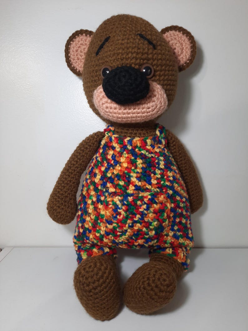 May include: A handmade brown crochet bear doll wearing colorful overalls. The bear has a black nose, pink muzzle, and light brown ears. The overalls are a mix of red, yellow, blue, and orange. The bear is sitting upright.