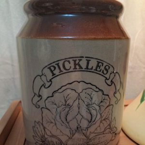 May include: A cylindrical stoneware crock with a brown glazed rim and a cork stopper. The body is light beige with a black line drawing of a cabbage and vegetables, featuring the word PICKLES printed in a banner above the illustration.