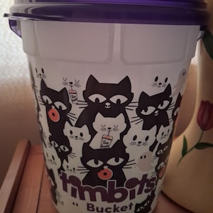 May include: A white plastic bucket with a purple lid featuring a repeating pattern of black and white cats, small ghosts, and Tim Hortons drink cups. The text "timbits Bucket" is printed in purple on the front of the container.