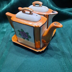 May include: A vintage ceramic teapot set in orange and white. The set comprises two teapots with lids and a shared tray. Each teapot has a floral design and a handle. A charming addition to any tea collection.