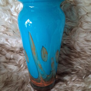 May include: A vibrant turquoise glass vase with a flared top. The vase is adorned with flowing orange and brown abstract patterns. The base of the vase is a darker brown colour. The vase is approximately 30 cm tall.