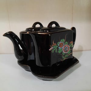 May include: Black ceramic teapot with a floral design in shades of orange, blue, and green. The teapot has a square shape with a spout, handle, and two lids. It sits on an attached rectangular base.