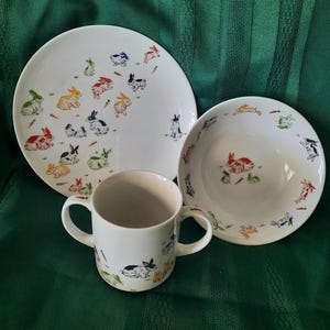Peut inclure: Un service de table en céramique blanche comprenant une assiette, un bol et une tasse. Chaque pièce est décorée d'illustrations colorées de lapins et de carottes. L'assiette est ronde, le bol est creux et la tasse a deux anses.