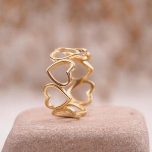 Peut inclure: Une bague dorée ornée de cœurs ajourés. La bague présente une série de cœurs découpés, créant un motif continu. Elle est présentée sur une surface neutre, mettant en valeur sa couleur dorée et son design détaillé.