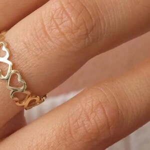 Pode incluir: Um anel dourado com um padrão de corações repetidos. O anel é usado em um dedo, mostrando o delicado design vazado dos corações. O tom quente do anel contrasta com o tom da pele, destacando o design da joia.