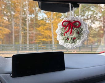 May include: A crocheted Christmas wreath car charm. The wreath is white and green with a red bow. The charm hangs from the rearview mirror in a car. The background shows autumn foliage.