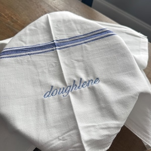 May include: A white kitchen towel featuring two horizontal blue stripes with a herringbone pattern and the name doughlene embroidered in blue script lettering across the center of the fabric.