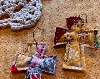 May include: A pair of cross-shaped earrings crafted from quilted fabric scraps. One earring features a yellow floral pattern with red accents, while the other displays a patchwork of various floral and checkered prints. Both have silver-toned metal hooks.