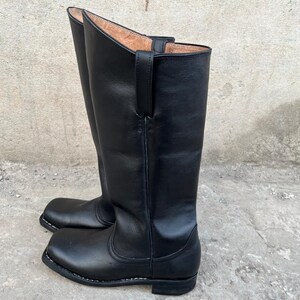 K&ouml;nnte beinhalten: Ein Paar hohe, schwarze Lederstiefel mit quadratischer Zehenpartie. Die Stiefel haben eine glatte, polierte Oberfl&auml;che und eine Schlaufe zum Anziehen oben. Die Sohlen sind schwarz mit wei&szlig;en N&auml;hten.