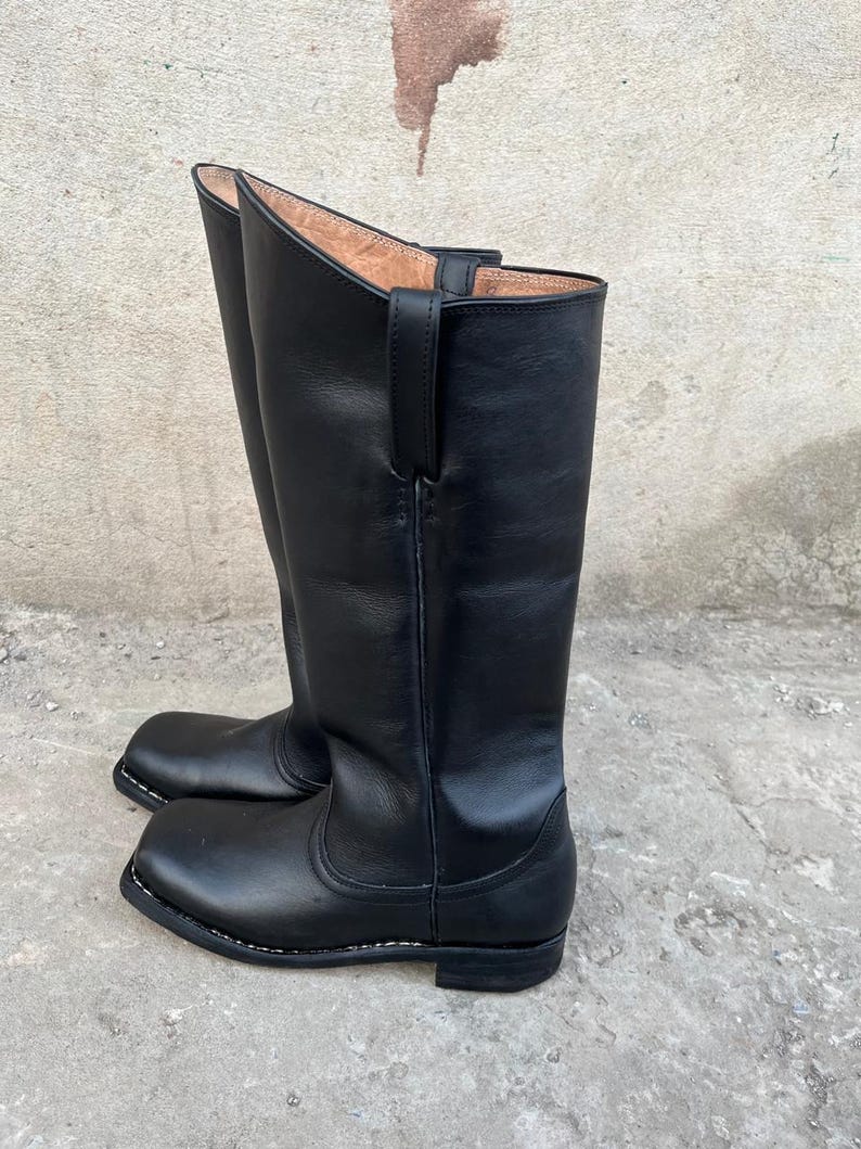 K&ouml;nnte beinhalten: Ein Paar hohe, schwarze Lederstiefel mit quadratischer Zehenpartie. Die Stiefel haben eine glatte, polierte Oberfl&auml;che und eine Schlaufe zum Anziehen oben. Die Sohlen sind schwarz mit wei&szlig;en N&auml;hten.