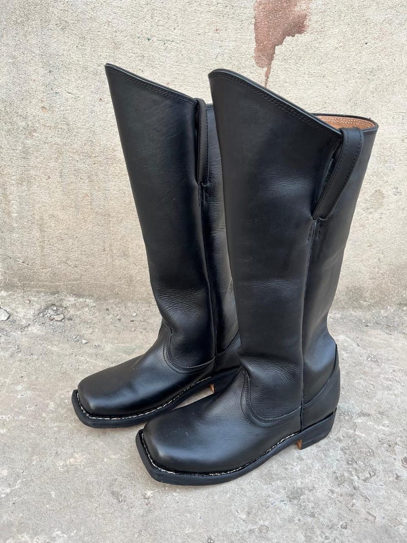 K&ouml;nnte beinhalten: Ein Paar schwarze Leder-Cowboystiefel. Die Stiefel haben einen hohen Schaft, eine quadratische Spitze und einen niedrigen Absatz. Die N&auml;hte sind wei&szlig;, und das Leder scheint glatt und poliert zu sein. Die Stiefel stehen auf einer Betonoberfl&auml;che.