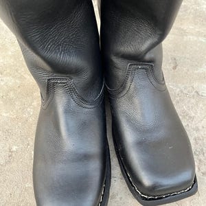 K&ouml;nnte beinhalten: Ein Paar schwarze Lederstiefel mit quadratischer Spitze. Die Stiefel haben eine glatte, gl&auml;nzende Oberfl&auml;che und eine gen&auml;hte Sohle. Die Sch&auml;fte der Stiefel erstrecken sich nach oben und zeigen einen klassischen, zeitlosen Stil.