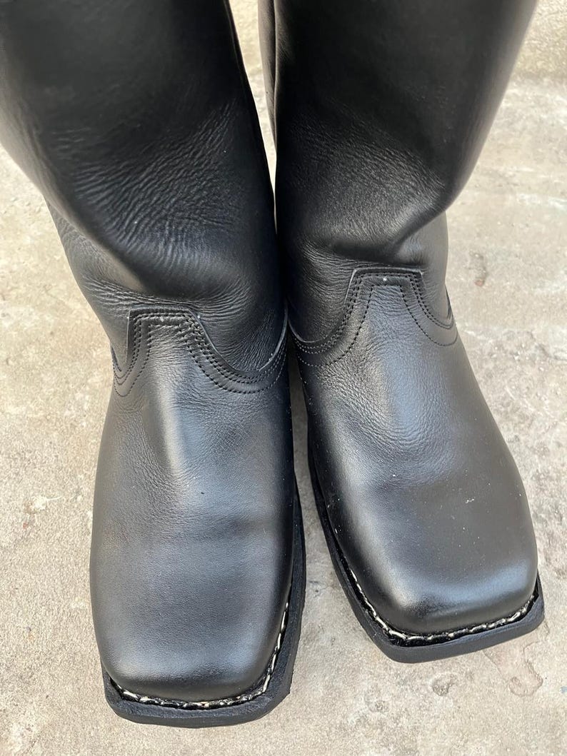 K&ouml;nnte beinhalten: Ein Paar schwarze Lederstiefel mit quadratischer Spitze. Die Stiefel haben eine glatte, gl&auml;nzende Oberfl&auml;che und eine gen&auml;hte Sohle. Die Sch&auml;fte der Stiefel erstrecken sich nach oben und zeigen einen klassischen, zeitlosen Stil.
