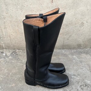 K&ouml;nnte beinhalten: Ein Paar schwarze Leder-Cowboystiefel. Die Stiefel sind hoch, mit abgerundeter Oberseite und quadratischer Spitze. Die N&auml;hte sind wei&szlig; und die Stiefel scheinen neu zu sein. Sie sind aus glattem, gl&auml;nzendem Leder gefertigt.