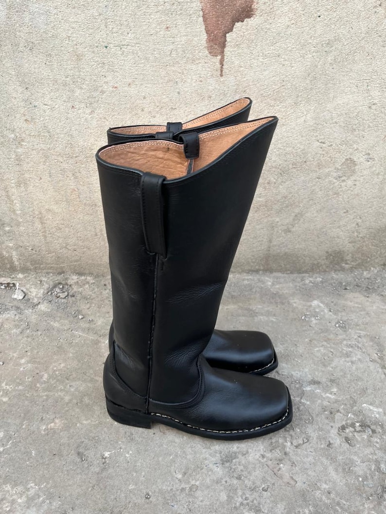 K&ouml;nnte beinhalten: Ein Paar schwarze Leder-Cowboystiefel. Die Stiefel sind hoch, mit abgerundeter Oberseite und quadratischer Spitze. Die N&auml;hte sind wei&szlig; und die Stiefel scheinen neu zu sein. Sie sind aus glattem, gl&auml;nzendem Leder gefertigt.