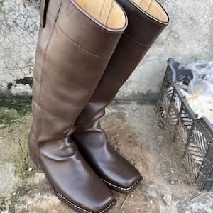May include: A pair of tall, brown leather riding boots. The boots have a square toe design with white stitching and a tall shaft. The leather appears smooth and the boots are standing upright.