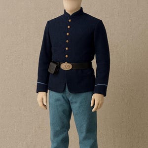 Könnte beinhalten: Eine marineblaue Uniform aus dem Bürgerkrieg auf einer Schaufensterpuppe. Die Uniform umfasst eine Jacke mit goldenen Knöpfen, einen passenden Hut und hellblaue Hosen. Ein schwarzer Gürtel mit Schnalle und eine kleine Tasche vervollständigen das Outfit.