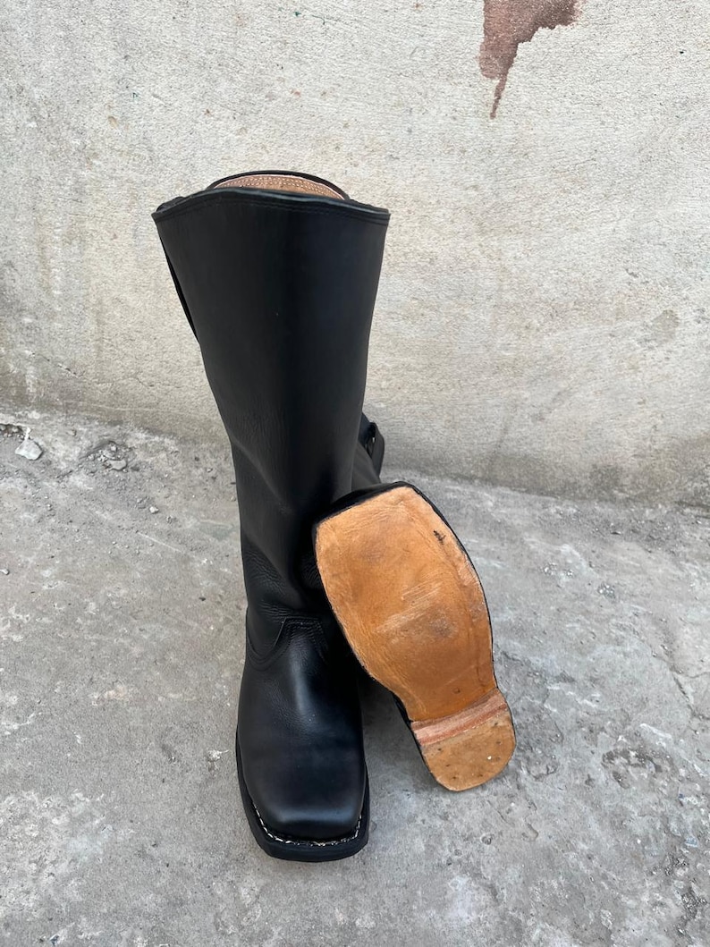 K&ouml;nnte beinhalten: Ein Paar schwarze Lederstiefel mit quadratischer Spitze und hohem Schaft. Ein Stiefel steht aufrecht, w&auml;hrend der andere auf der Seite liegt und die hellbraune Sohle freilegt. Die Stiefel scheinen neu zu sein.