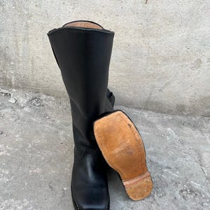 K&ouml;nnte beinhalten: Schwarze Lederreitstiefel mit quadratischer Spitze und hohem Schaft. Die Sohle ist sichtbar und hat eine hellbraune Farbe. Die Stiefel stehen auf einer Betonoberfl&auml;che.