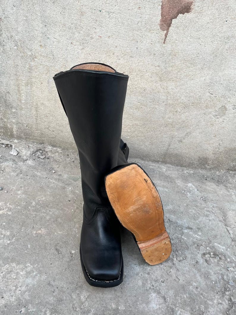 K&ouml;nnte beinhalten: Schwarze Lederreitstiefel mit quadratischer Spitze und hohem Schaft. Die Sohle ist sichtbar und hat eine hellbraune Farbe. Die Stiefel stehen auf einer Betonoberfl&auml;che.