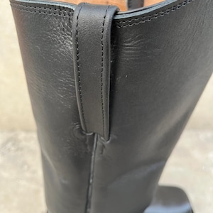K&ouml;nnte beinhalten: Nahaufnahme eines schwarzen Lederstiefels. Der Stiefel hat eine Zuglasche oben und sichtbare N&auml;hte. Das Leder wirkt glatt und der Stiefel hat ein klassisches Design. Der Fokus liegt auf dem oberen Teil des Stiefels.