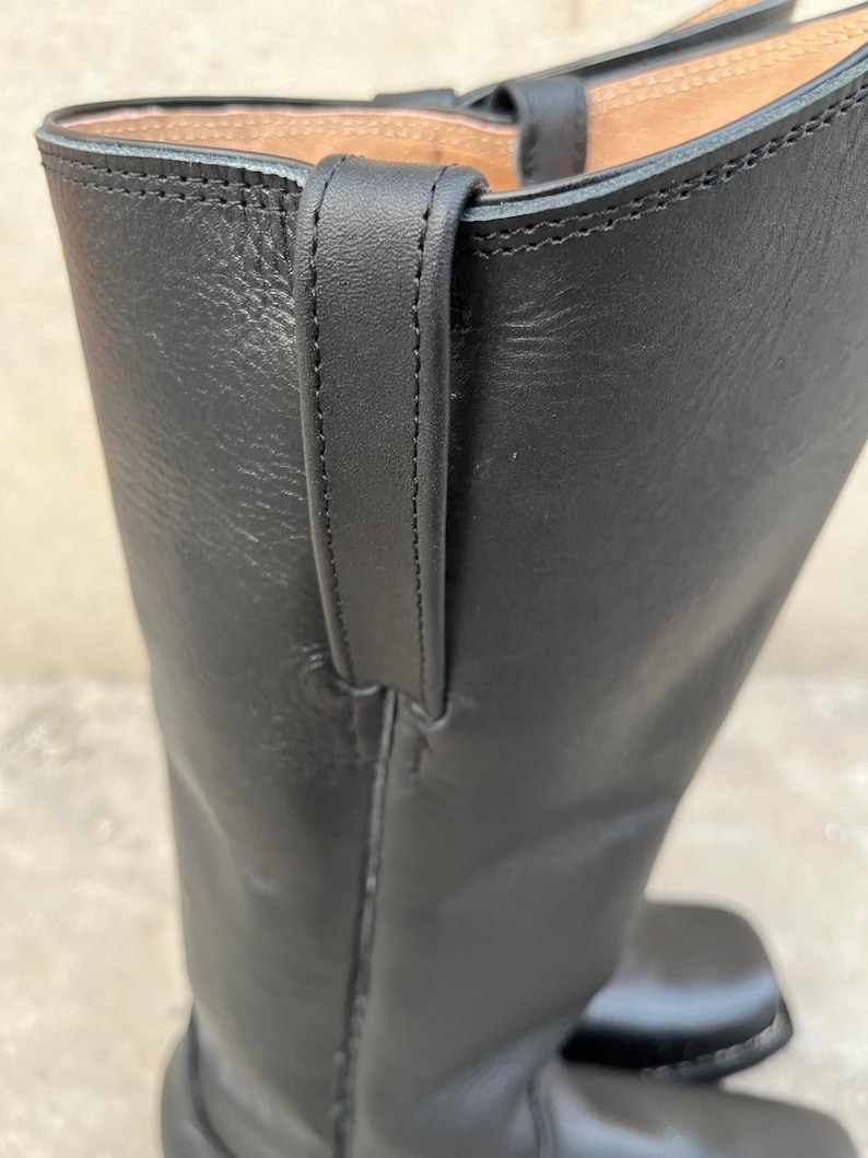 K&ouml;nnte beinhalten: Nahaufnahme eines schwarzen Lederstiefels. Der Stiefel hat eine Zuglasche oben und sichtbare N&auml;hte. Das Leder wirkt glatt und der Stiefel hat ein klassisches Design. Der Fokus liegt auf dem oberen Teil des Stiefels.