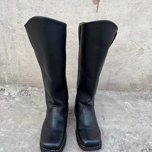 K&ouml;nnte beinhalten: Ein Paar schwarze Kniehochstiefel aus Leder. Die Stiefel haben eine quadratische Zehenpartie und eine wei&szlig; gen&auml;hte Sohle. Die Stiefel stehen auf einer Betonoberfl&auml;che.