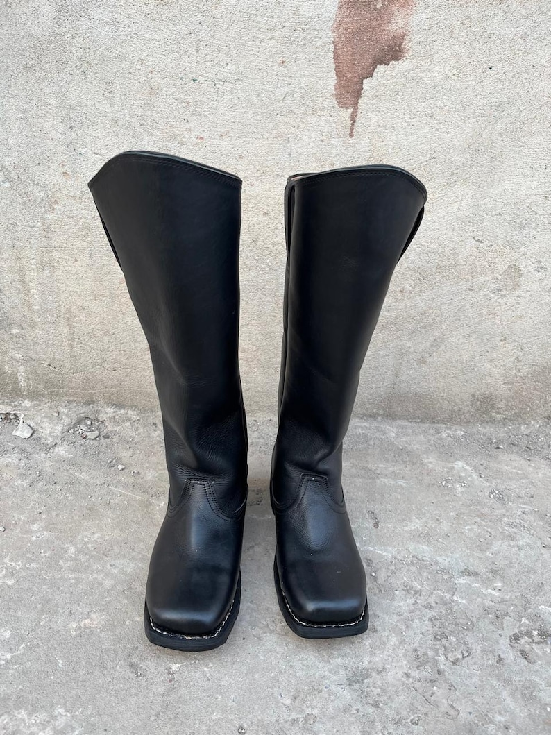 K&ouml;nnte beinhalten: Ein Paar schwarze Kniehochstiefel aus Leder. Die Stiefel haben eine quadratische Zehenpartie und eine wei&szlig; gen&auml;hte Sohle. Die Stiefel stehen auf einer Betonoberfl&auml;che.