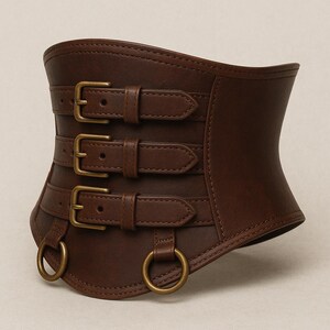 May include: A brown leather corset belt with three buckle straps and two metal rings. The belt is designed to cinch the waist and has a curved shape. The leather has a textured appearance.
