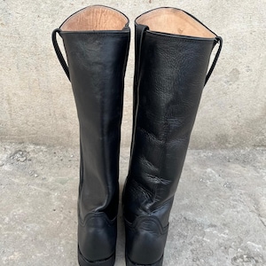 K&ouml;nnte beinhalten: Ein Paar schwarze Lederreitstiefel. Die Stiefel sind hoch, reichen bis zur Mitte der Wade und haben eine glatte, polierte Oberfl&auml;che. Sie haben ein klassisches Design mit runder Spitze und kleinem Absatz. Das Innenfutter ist hellbraun.