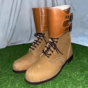 May include: A pair of tan suede boots with brown laces and a brown leather cuff secured by buckles. The boots have a round toe and a dark brown sole. The boots are on a green surface.