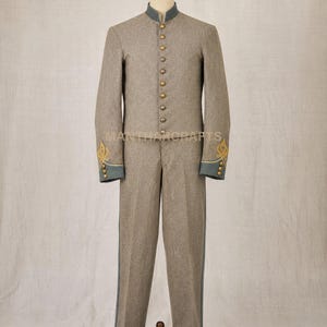 Può includere: Un completo uniforme in misto lana beige composto da una giacca a maniche lunghe e pantaloni abbinati. La giacca presenta un colletto alto, una fila verticale di bottoni in ottone e polsini blu con ricami dorati. Il testo MANTHACRAFTS è visibile.