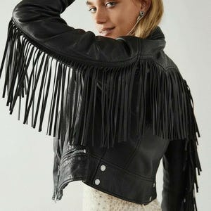 May include: Black leather jacket with long fringe detailing across the shoulders and sleeves. The jacket has a zip closure and two press studs. The model is wearing a white lace dress.