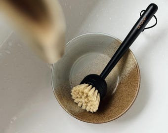 May include: A black handled dish brush with natural bristles rests inside a ceramic bowl. The bowl has a brown and beige speckled design. The brush handle has the text "DETOORIP" printed on it.
